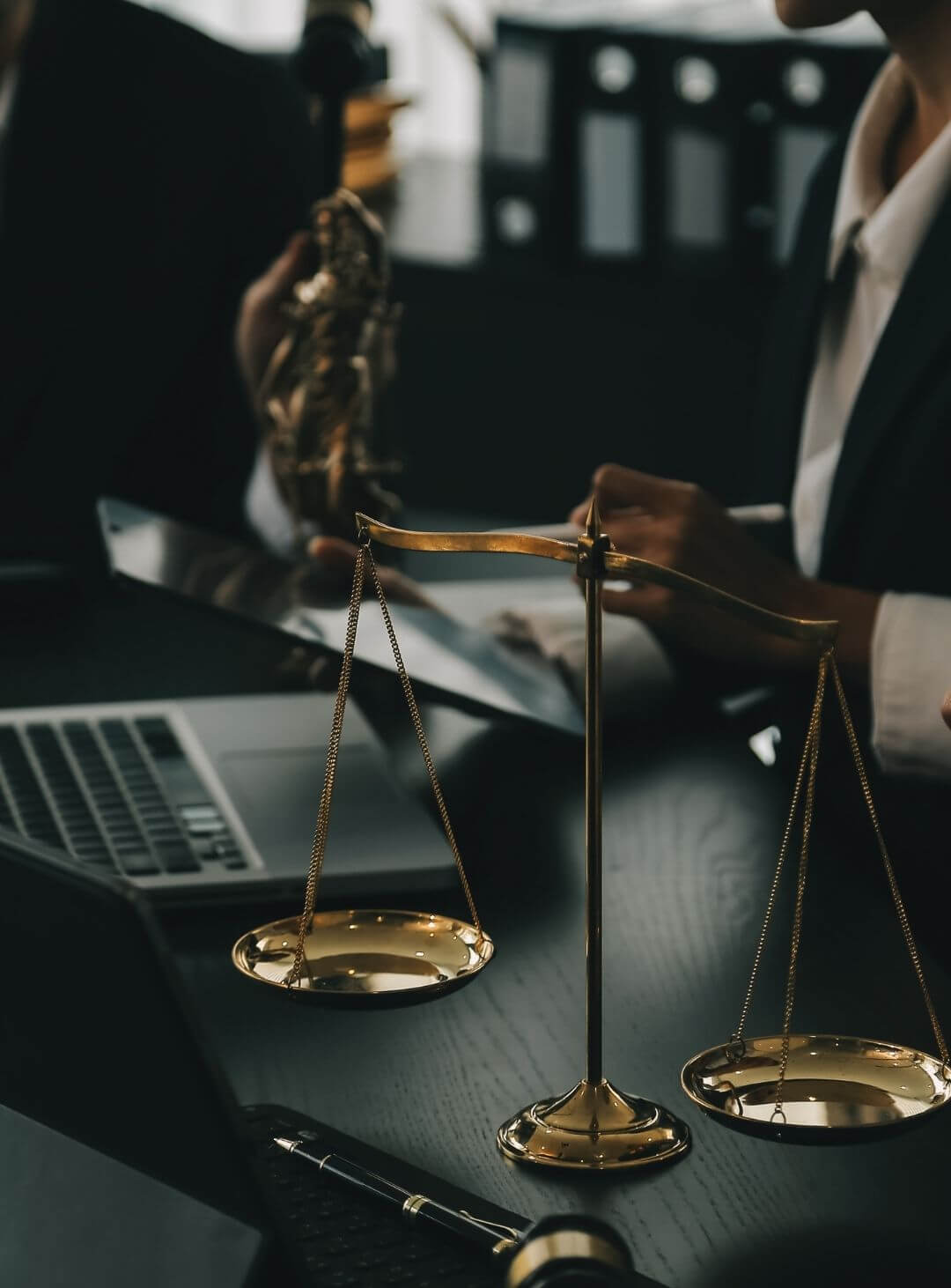 Close-up de uma balança da justiça dourada sobre uma mesa de escritório, com advogado e laptop ao fundo, simbolizando a solidez jurídica, o equilíbrio e a segurança oferecidos pela Fernanda Souza Advocacia.